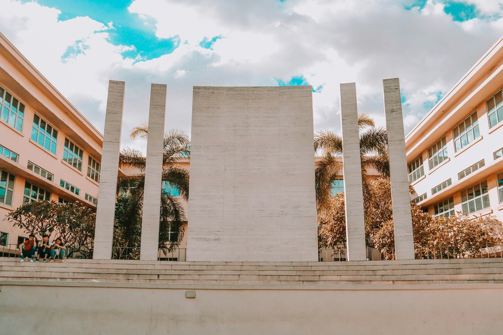A wide plaza between two school buildings on a Philippine campus, with students sitting together in the shade
